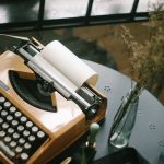 Een vintage Olivetti typemachine op een bureau naast een vaas met bloemen.