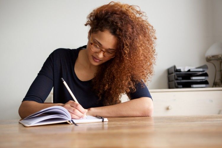 Een schrijver met krullend haar die geconcentreerd in een notitieboek schrijft.